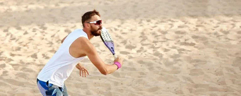 Dynamic image of young man playing beach tennis, hitting ball with racket. Outdoor training on warm summer day. Top view. Concept of sport, leisure time, active lifestyle, hobby, game, summertime, ad